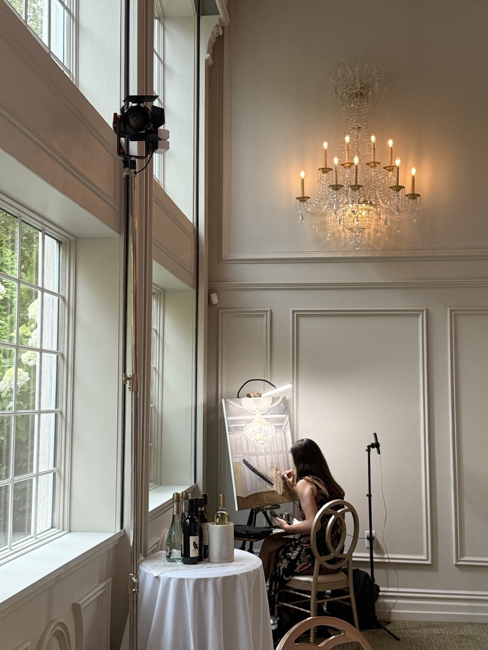 Live wedding painter creates a custom portrait during an elegant reception at Florentine Gardens in River Vale, New Jersey, with crystal sconces, tall windows, and a softly lit ballroom in the background.